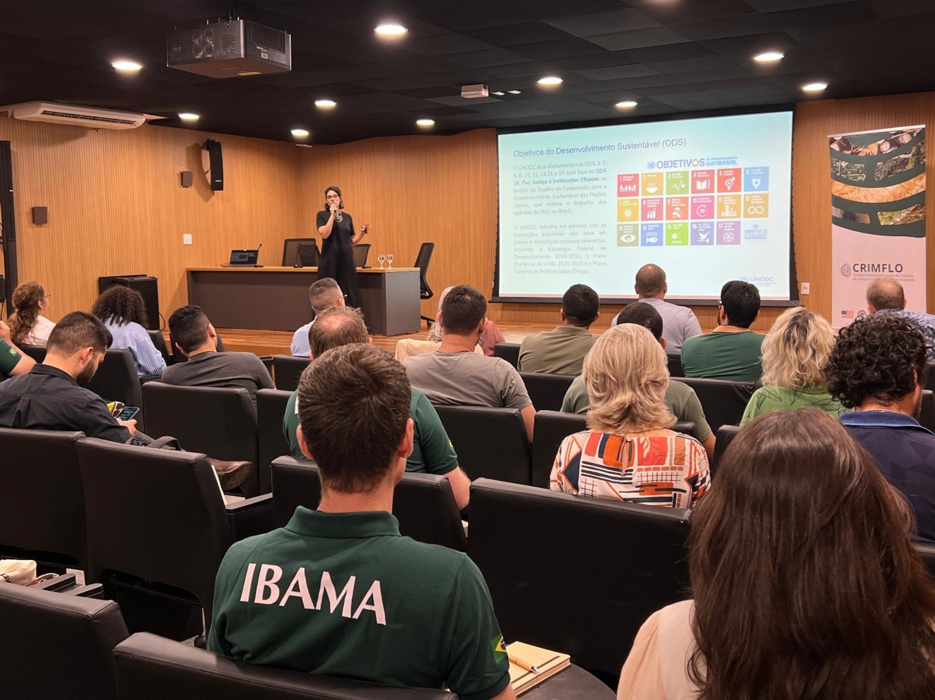 Nesta imagem, vemos uma apresentação em um auditório. Uma pessoa, à frente, está falando ao microfone e projetando uma tela com informações sobre os Objetivos de Desenvolvimento Sustentável (ODS) da ONU, com destaque para os ícones coloridos dos 17 objetivos. Na plateia, pessoas estão sentadas, muitas delas vestindo camisetas com o logo do IBAMA, voltadas para a palestrante. O ambiente tem iluminação de teto discreta e design moderno, com painéis de madeira nas paredes e cadeiras pretas. À direita, um banner com o logo do CRIMFLO está visível.